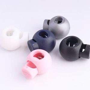 May include: Five plastic toggle cord locks in different colors: white, navy blue, black, silver, and pink. Each toggle has a spring inside and a hole for threading cord.