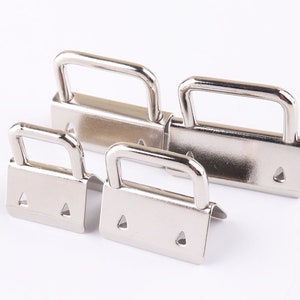May include: Four silver metal slider buckles of different sizes. The buckles have a rectangular shape with a D-shaped loop on top. The buckles are commonly used for straps, bags, and clothing.