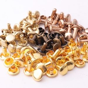 Op de afbeelding: Een verzameling metalen klinknagels in verschillende kleuren, waaronder goud, brons en donkerbrons. De klinknagels hebben een afgeronde kop en een pen voor bevestiging. Deze bevestigingsmiddelen worden vaak gebruikt in leerbewerking en kledingfabricage.