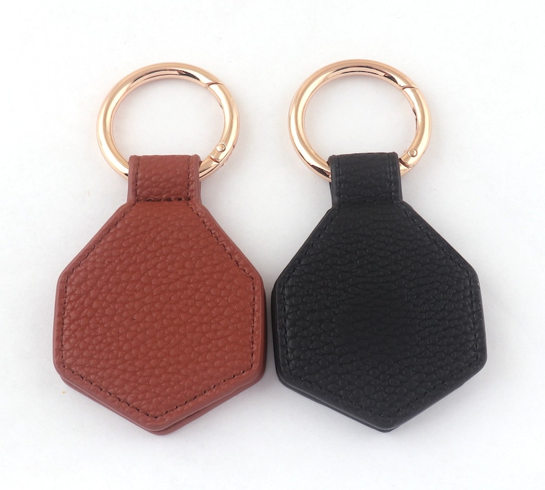 May include: Two leather keychains with hexagonal shapes. One is brown and the other is black. Both have gold metal rings.
