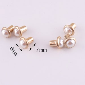 Puede incluir: Seis tachuelas de metal dorado con tapas de perla blanca. Las tachuelas tienen un di&aacute;metro de 6 mm y 7 mm.