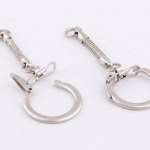 Puede incluir: Dos llaveros plateados con un cordón retráctil de resorte y un anillo circular. Los anillos están abiertos y tienen un clip en el lateral.