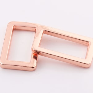 Può includere: Due fibbie rettangolari scorrevoli in metallo color oro rosa. Le fibbie sono impilate l'una sull'altra.
