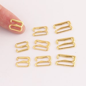 Pode incluir: Um conjunto de dez ajustadores de alça de sutiã de metal dourado. Os ajustadores são de vários tamanhos e estão dispostos em uma fila sobre uma superfície branca.
