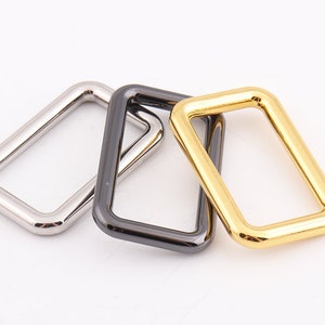 May include: Three metal rectangular rings in silver, black, and gold. These hardware components are likely used in bag making, jewelry, or other craft projects. The rings are displayed on a white background.