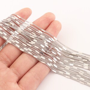 May include: A silver chain necklace with a delicate, flat, rectangular link design. The chain is shown in a pile, with multiple strands layered on top of each other.