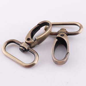 May include: Two antique gold metal swivel clasps, each with a D-ring and a hook. The clasps are designed for attaching to straps, bags, or other items.