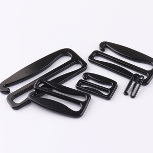 May include: A set of five black metal bra strap adjusters. The adjusters are in various shapes and sizes, with some having a hook and others having a loop.