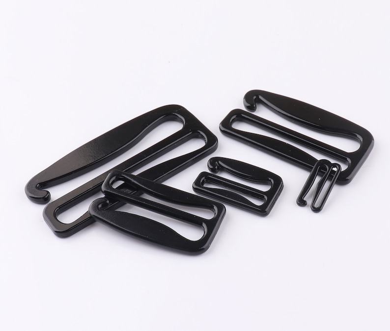 May include: A set of five black metal bra strap adjusters. The adjusters are in various shapes and sizes, with some having a hook and others having a loop.