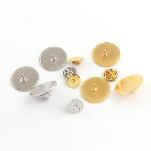May include: A collection of various metal pin backs in gold and silver tones. The pin backs are round and oval, with different styles of clasps and posts. Some have a flat back, while others have a textured surface.