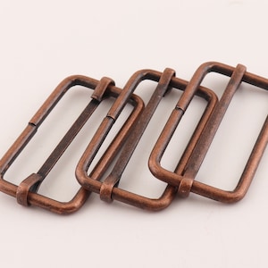 May include: Three antique copper-colored metal slider buckles. The buckles are rectangular with rounded corners and have a smooth, slightly textured surface.