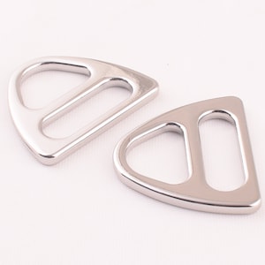 Op de afbeelding: Twee zilveren metalen D-ring gespen. De gespen zijn glad en glanzend. Ze zijn beide scherp en liggen op een wit oppervlak.