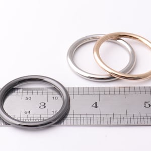 Puede incluir: Tres anillos metálicos en plata, dorado y gris oscuro se muestran sobre una superficie blanca junto a una regla. El anillo gris oscuro tiene aproximadamente 7,6 cm de diámetro. Los otros dos anillos son de tamaño similar.