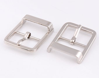 May include: Two silver metal belt buckles. The buckles are rectangular with a smooth, polished finish. The buckles are designed to be used with a belt strap.