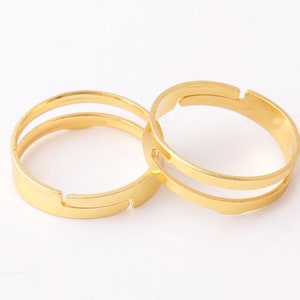 Op de afbeelding: Twee gouden metalen ringblanken met een dubbele ringontwerp. De ringen zijn aan één kant open en kunnen worden gebruikt om aangepaste ringen te maken.