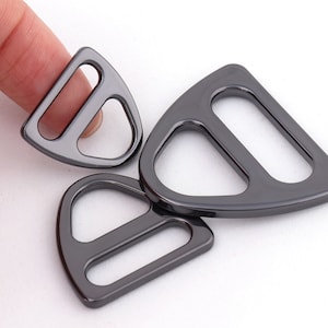 Könnte beinhalten: Drei schwarze D-Ringe aus Metall. Die Ringe haben die Form eines Dreiecks mit abgerundeten Ecken. Die Ringe werden zum Befestigen von Bändern oder anderen Materialien verwendet.
