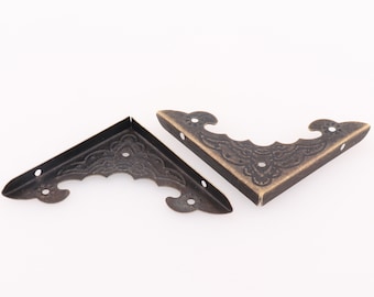 Può includere: Due staffe d'angolo in ottone antico con un design ornamentale. Le staffe sono in metallo e hanno una finitura marrone scuro.