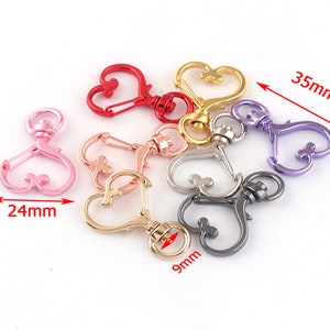 Könnte beinhalten: Sortiment aus sechs herzförmigen Schlüsselanhängern aus Metall in verschiedenen Farben, darunter Rot, Rosa, Gold, Silber und Lila. Jeder Schlüsselanhänger ist etwa 3,5 cm lang und verfügt über einen drehbaren Karabinerhaken.