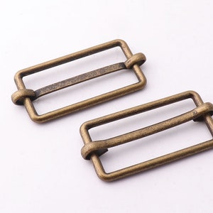 Könnte beinhalten: Zwei antike, goldfarbene Metall-Schiebeschließen. Die Schließen sind rechteckig mit abgerundeten Ecken und haben einen einzigen Steg in der Mitte.