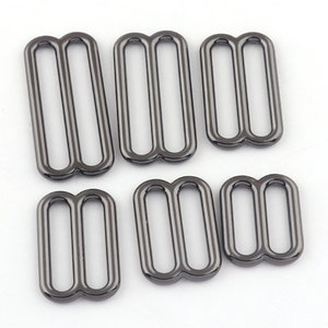 May include: Six silver-toned metal slider buckles of varying sizes. The buckles have a double loop design and are commonly used for adjusting straps on clothing or bags.