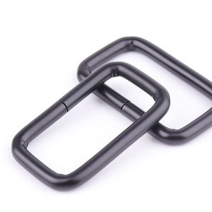May include: Two black metal D-rings, one slightly larger than the other, are stacked on top of each other. The rings are smooth and have a matte finish.