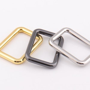 May include: Three metal D-rings in gold, black, and silver. The rings are square shaped and have a smooth finish.