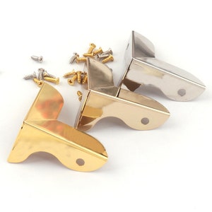 May include: Three corner brackets in gold, silver, and nickel finishes. The brackets are designed to reinforce corners of furniture or boxes. The brackets are shown on a white background with screws.