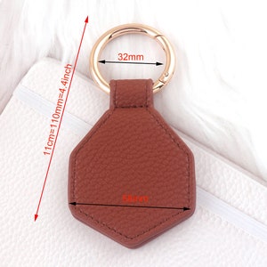 May include: A brown leather hexagon-shaped keychain with a gold metal ring. The keychain measures 5.8 cm wide and 11 cm long.