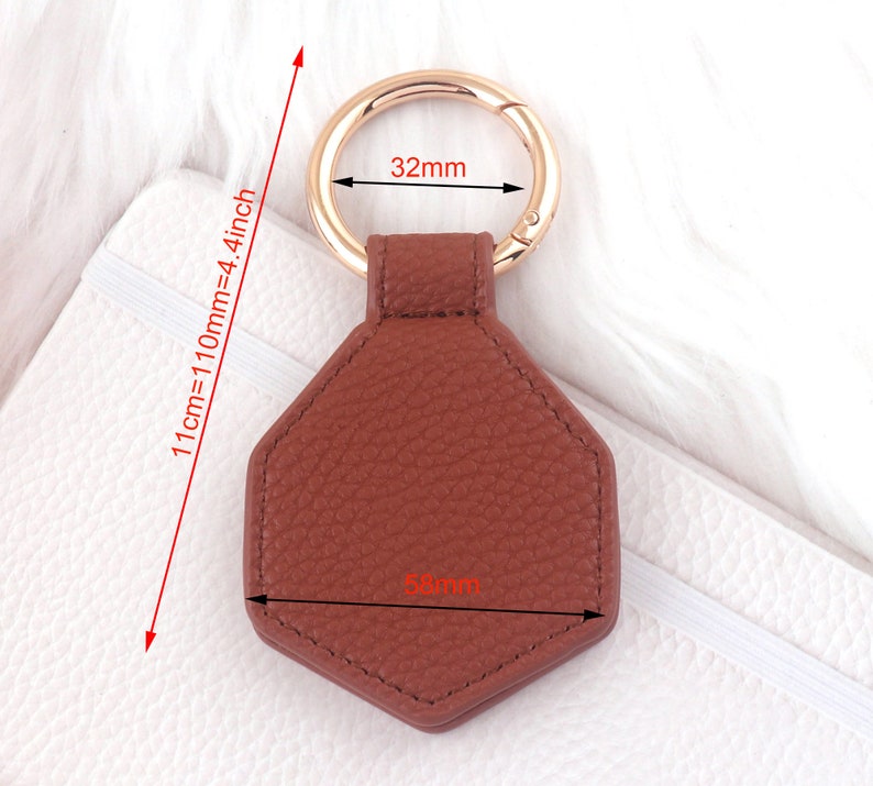 May include: A brown leather hexagon-shaped keychain with a gold metal ring. The keychain measures 5.8 cm wide and 11 cm long.
