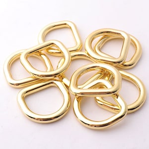 Op de afbeelding: Een groep gouden metalen D-ringen, ook wel D-ringen of D-vormige ringen genoemd. Deze ringen worden vaak gebruikt om banden, kettingen of andere hardware aan tassen, riemen of andere accessoires te bevestigen.