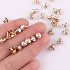 Puede incluir: Una colecci&oacute;n de tachuelas de metal dorado con tapas de color blanco perlado. Las tachuelas est&aacute;n esparcidas sobre una superficie blanca.