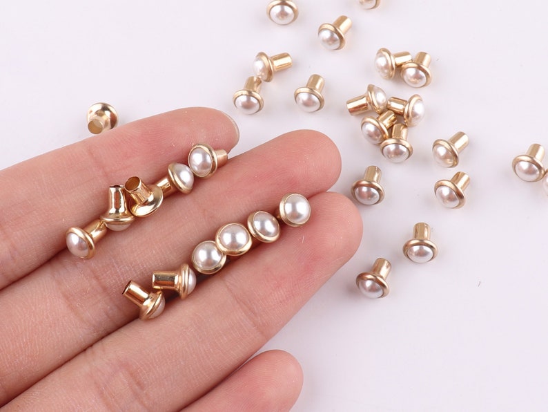Puede incluir: Una colecci&oacute;n de tachuelas de metal dorado con tapas de color blanco perlado. Las tachuelas est&aacute;n esparcidas sobre una superficie blanca.