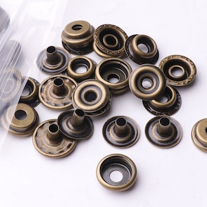 May include: A close-up of a pile of antique gold metal snap fasteners. The fasteners are round and have a raised centre with a hole in the middle. The fasteners are scattered on a white surface.