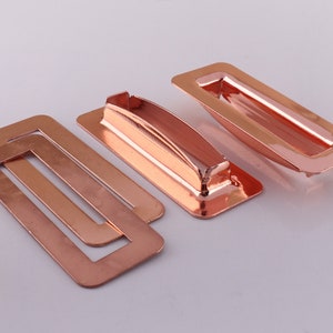 Könnte beinhalten: Drei roségoldene Schubladengriffe, zwei sind rechteckig mit einem Rand und einer ist ein rechteckiger Griff mit einem erhöhten Mittelteil.