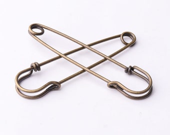 May include: Two antique bronze safety pins crossed against a white background. The pins have a classic design with a curved bottom and a coiled top, showcasing a vintage aesthetic. Ideal for craft projects or adding a unique touch to clothing.