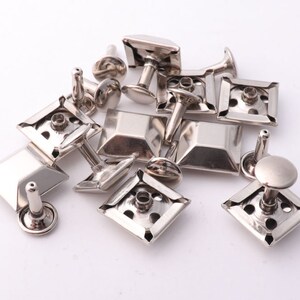 May include: A collection of silver-toned metal rivets with square and rectangular heads. The rivets have a post that can be hammered or pressed into place.