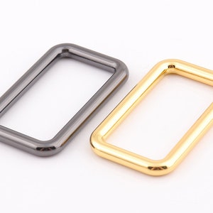 May include: Two rectangular metal rings. One is a dark gunmetal gray, the other is a shiny gold color. The rings are likely used for bag hardware or other crafting projects. The shapes are smooth and rounded.