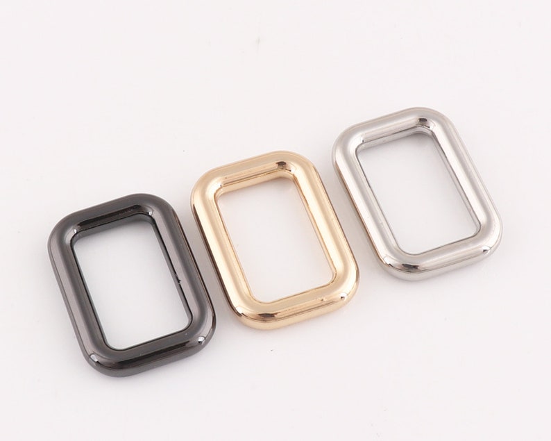 Puede incluir: Tres anillos rectangulares de metal en diferentes acabados: negro, dorado y plateado. Los anillos miden aproximadamente 5 cm de largo y 2,5 cm de ancho.
