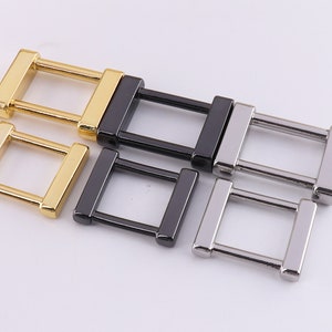 May include: Four metal belt buckle loops in gold, black, and silver. The loops are rectangular with a square opening in the center.