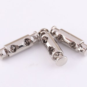 May include: Three silver metal cord stoppers with a spring mechanism. The stoppers are designed to hold cords or straps in place.