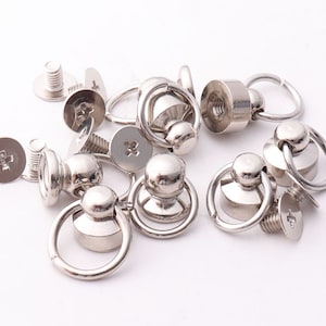 Könnte beinhalten: Nahaufnahme von silbernen Drehschnapphaken aus Metall mit einem runden Ring. Die Haken sind zum Befestigen an Stoff oder Leder gedacht.