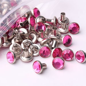 Puede incluir: Una bolsa de tachuelas de metal con base plateada y adorno de strass rosa. Las tachuelas están esparcidas sobre una superficie blanca.