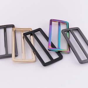 May include: Five metal belt buckle slides in different finishes. The slides are rectangular with a single, horizontal slot in the center. The finishes include black, gold, and iridescent.