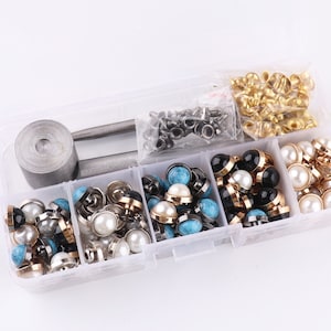 Op de afbeelding: Een transparante plastic opbergdoos gevuld met verschillende soorten metalen en plastic knopen. De doos bevat een gereedschap voor het bevestigen van de knopen. De knopen zijn in verschillende kleuren, waaronder goud, zilver, zwart, wit en turquoise.