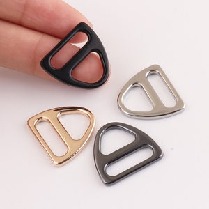 Pode incluir: Quatro fivelas de metal em forma de D em diferentes acabamentos: preto, prata, dourado e bronze. As fivelas têm a forma de uma lágrima com a parte superior arredondada e a inferior plana.