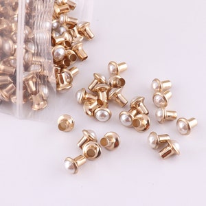 Puede incluir: Una bolsa de tachuelas de metal dorado con tapas de perlas blancas. Las tachuelas est&aacute;n esparcidas sobre una superficie blanca.