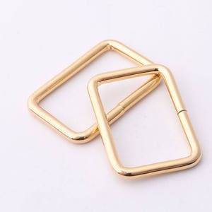 May include: Two gold-colored metal D-rings, one slightly larger than the other, are shown on a white background.