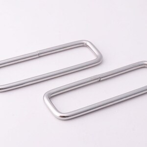 May include: Two silver-colored, rectangular metal rings. Each ring is a continuous loop with a slight gap on one side. The rings are smooth and have rounded corners, suggesting a minimalist design.