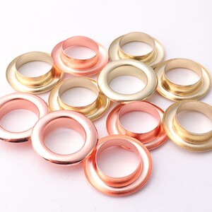 Op de afbeelding: Een verzameling van gouden en roségouden metalen oogjes. De oogjes zijn willekeurig gerangschikt op een wit oppervlak.