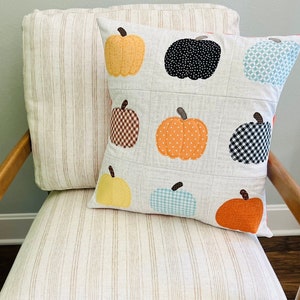 Pode incluir: Uma almofada branca com um design de patchwork que apresenta várias abóboras de cores e padrões. A almofada é feita de tecido e tem uma textura macia e confortável.